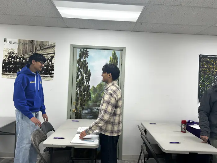 Two people talking beside a table in a classroom before the competition starts.